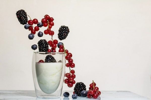 Kapil Abhijat Product 3, a double-walled glass filled with white yogurt, is topped with blackberries, blueberries, and red currants. While some berries appear to be falling into the yogurt, others are scattered around the base of the glass on a white surface.